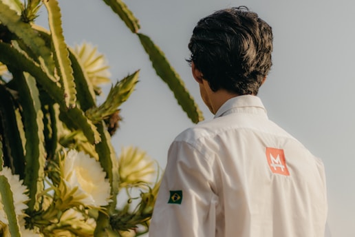 A person with dark hair is wearing a white shirt with a logo on the back and a small Brazilian flag on the sleeve, standing near tall cacti with large white flowers.
