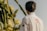 A person with dark hair is wearing a white shirt with a logo on the back and a small Brazilian flag on the sleeve, standing near tall cacti with large white flowers.