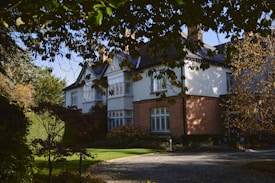 A large, classic-style house surrounded by lush greenery, with a red-brick and white exterior. Leafy branches frame the view, casting shadows over the garden and driveway. The scene is tranquil and bathed in natural light.