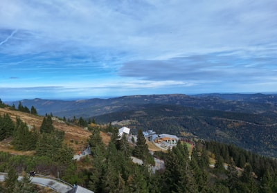 A vast landscape featuring a mountainous terrain covered with dense pine forests. A winding road leads to a modern building with long pathways. The sky is expansive with patches of blue intermingled with clouds, providing a sense of openness.