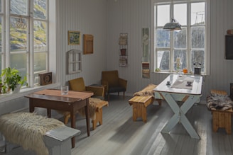 A cozy room with wooden floors and white wooden walls. There are several pieces of vintage furniture, including a table with a glass and a bench covered in fur throws. The room features large windows that let in natural light and offer a view of an outdoor landscape with a body of water and grassy hills. Various decorations such as paintings, shelves, and a radiator are mounted on the walls. Plants and small items like mugs and lamps are placed on the tables.