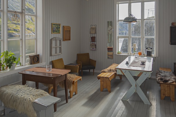 A cozy room with wooden floors and white wooden walls. There are several pieces of vintage furniture, including a table with a glass and a bench covered in fur throws. The room features large windows that let in natural light and offer a view of an outdoor landscape with a body of water and grassy hills. Various decorations such as paintings, shelves, and a radiator are mounted on the walls. Plants and small items like mugs and lamps are placed on the tables.