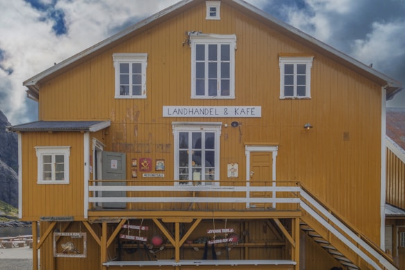 A rustic wooden building with a mustard yellow facade features a sign indicating it's a combination of a general store and café. The structure includes several windows with white frames, a small porch, and an exterior staircase leading to an upper entrance. Various signs and items, including a post box and advertisement signs for tours, can be seen along the wall.