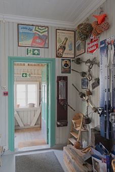 A quaint interior space with a vintage touch, featuring wooden paneled walls and a collection of old-fashioned advertisements and signs. There is a doorway leading to another room with natural light coming from a window. Various nautical-themed decorations and fishing rods are organized on the wall, along with a small shelf holding multiple items. The color scheme includes light pastel tones with turquoise accents on the door frame.