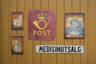 A wooden wall displays vintage signs, including a central red sign with a postal horn and the word 'POST' beneath a crown. On either side are rusty, illustrated metal signs featuring nostalgic imagery. Below, a sign reads 'MEDISINUTSALG' in bold black letters on a white background.