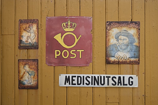 A wooden wall displays vintage signs, including a central red sign with a postal horn and the word 'POST' beneath a crown. On either side are rusty, illustrated metal signs featuring nostalgic imagery. Below, a sign reads 'MEDISINUTSALG' in bold black letters on a white background.