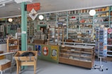 A vintage general store interior with a variety of goods displayed on shelves, including canned food, scales, and old-fashioned signs. The store features a wooden floor, teal and cream-painted shelves, and a rustic ambiance. An old cash register and weighing scale are present on the counter.
