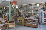 A vintage general store interior with a variety of goods displayed on shelves, including canned food, scales, and old-fashioned signs. The store features a wooden floor, teal and cream-painted shelves, and a rustic ambiance. An old cash register and weighing scale are present on the counter.