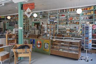 A vintage general store interior with a variety of goods displayed on shelves, including canned food, scales, and old-fashioned signs. The store features a wooden floor, teal and cream-painted shelves, and a rustic ambiance. An old cash register and weighing scale are present on the counter.