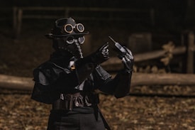 A person dressed in a dark plague doctor costume, complete with a beak-like mask, goggles, and a wide-brimmed hat, is standing outdoors. They are holding a syringe in one hand and appear to be examining it closely. The background is dimly lit, and the wooden fencing and sparse foliage suggest an eerie, historical atmosphere.