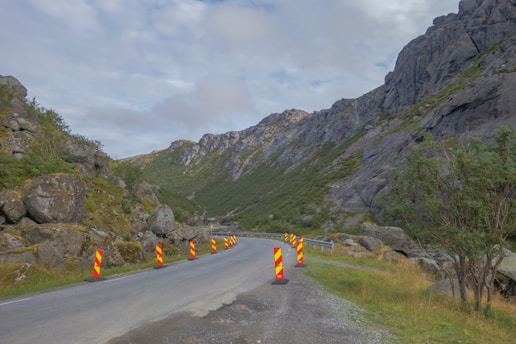 A winding road passes through a mountainous landscape, flanked by rocky cliffs and green vegetation. Orange and yellow traffic bollards line the road, indicating construction or maintenance work. The sky is overcast with clouds, creating a subdued atmosphere.