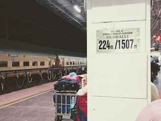 A train station platform with a marked sign indicating it is Hubballi, the world's longest platform at 224 meters or 1507 meters. There is a stationary train on the tracks and a cart loaded with luggage in the foreground. The setting appears to be at night, with artificial lighting illuminating the platform.