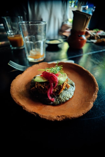 A rustic plate holds a vibrant dish featuring a blue corn tortilla topped with shredded meat, onions, avocado, and garnished with a sprig of fresh herbs. In the background, there are two glasses, one of which is partially filled, and a small clay jug, all set on a dark, reflective surface.