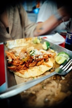 A deliciously presented taco featuring marinated meat garnished with onions and herbs. The taco sits on a corn tortilla, accompanied by sliced limes on a plate. A fork rests nearby, suggesting the dish is about to be enjoyed. The meal is set against a casual dining environment with a blurred background.