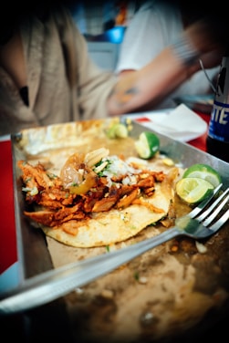 A deliciously presented taco featuring marinated meat garnished with onions and herbs. The taco sits on a corn tortilla, accompanied by sliced limes on a plate. A fork rests nearby, suggesting the dish is about to be enjoyed. The meal is set against a casual dining environment with a blurred background.