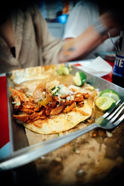 A deliciously presented taco featuring marinated meat garnished with onions and herbs. The taco sits on a corn tortilla, accompanied by sliced limes on a plate. A fork rests nearby, suggesting the dish is about to be enjoyed. The meal is set against a casual dining environment with a blurred background.