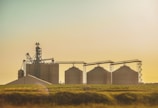 Several large grain silos are set against a golden sky. The silos are accompanied by agricultural equipment and are surrounded by fields, indicating a rural agricultural setting.