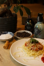 A plate of spaghetti topped with tomato sauce, grated cheese, and garnished with parsley sits on a table. Nearby, there is a cork, a bowl of salt, a bowl of peppercorns, and bottles of balsamic vinegar and olive oil. A potted plant is in the background.