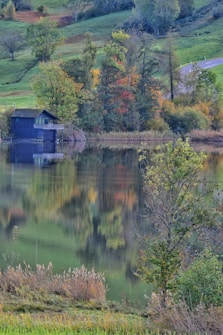 A serene lakeside scene featuring a small wooden cabin surrounded by lush greenery and colorful autumnal trees. The reflections on the calm water create a mirror image of the landscape. A winding road is visible in the background, adding depth to the picturesque setting.