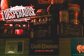 A dimly lit bar or pub interior with various vintage-style beer and liquor advertisements on wooden crates. Prominent brands like Desperados and Voll-Damm are visible, along with other labels. A no-smoking sign is displayed as well. The overall setting is rustic and nostalgic.