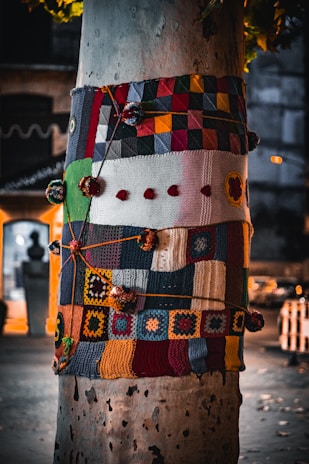 A tree trunk wrapped in a colorful patchwork of knitted and crocheted squares and patterns. The yarn artwork features an array of geometric shapes, vibrant colors, and varied textures, decorated with pom-poms and strings.