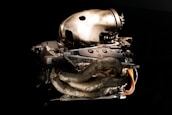 A high-performance car engine featuring intricate carbon fiber components and metallic parts is set against a dark background. The engine's design is modern and aerodynamic, showcasing exposed wiring and tubes.