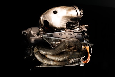 A high-performance car engine featuring intricate carbon fiber components and metallic parts is set against a dark background. The engine's design is modern and aerodynamic, showcasing exposed wiring and tubes.