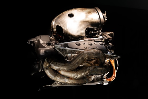 A high-performance car engine featuring intricate carbon fiber components and metallic parts is set against a dark background. The engine's design is modern and aerodynamic, showcasing exposed wiring and tubes.