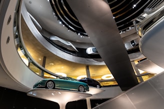 An indoor multi-level display of various cars showcased in a modern, futuristic setting. The display involves circular platforms that hold the cars at different levels, with a metallic spiral ramp intertwining through the space. The area is well-lit with bright lights from the ceiling.