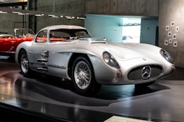 A sleek, vintage sports car displayed in a museum setting. The car features a shiny silver exterior, classic aerodynamic design, and distinctive wire-spoke wheels. It has a low profile and a prominent front grille with an iconic emblem. The background includes a red car and a modern, minimalist interior with concrete walls and soft lighting.
