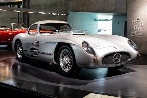 A sleek, vintage sports car displayed in a museum setting. The car features a shiny silver exterior, classic aerodynamic design, and distinctive wire-spoke wheels. It has a low profile and a prominent front grille with an iconic emblem. The background includes a red car and a modern, minimalist interior with concrete walls and soft lighting.