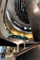 Interior view of a modern, multi-level showroom with sleek architectural design elements. A green sports car is prominently displayed on a raised white platform, surrounded by floors displaying other vehicles. The overhead lighting and spiral design contribute to a futuristic ambiance.