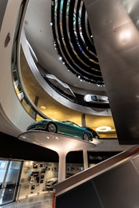 Interior view of a modern, multi-level showroom with sleek architectural design elements. A green sports car is prominently displayed on a raised white platform, surrounded by floors displaying other vehicles. The overhead lighting and spiral design contribute to a futuristic ambiance.