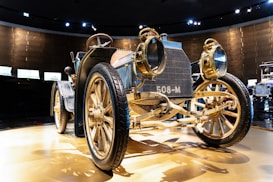 A vintage automobile with brass details and large, spoked wheels is displayed prominently in a well-lit room. Its design reflects an early 20th-century style, with a prominent grill marked '508-M'. The car's body is composed of a mix of metal and wood elements, showcasing intricate craftsmanship. The setting suggests this is part of a collection in a museum or exhibition space.