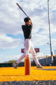 A glass bottle of apple soda is prominently positioned on a vivid yellow surface, with a blurred baseball player in the background preparing to swing a bat. The sky is partly cloudy, adding a dynamic backdrop to the scene.