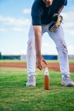 A person wearing a baseball glove is reaching towards a glass bottle of Manzana soda placed on a grassy field. The individual is dressed in athletic attire, including a navy blue shirt and white pants. The background features a blurred view of the baseball field with a clear sky overhead.