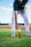 A person wearing a baseball glove is reaching towards a glass bottle of Manzana soda placed on a grassy field. The individual is dressed in athletic attire, including a navy blue shirt and white pants. The background features a blurred view of the baseball field with a clear sky overhead.