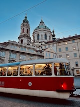 A red and white tram is filled with passengers, moving through a historic cityscape. In the background, a grand, ornate church with a domed roof and two towers stands under a clear blue sky. The scene conveys a blend of modern urban life and classic architectural beauty.