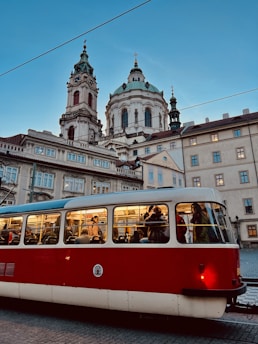 A red and white tram is filled with passengers, moving through a historic cityscape. In the background, a grand, ornate church with a domed roof and two towers stands under a clear blue sky. The scene conveys a blend of modern urban life and classic architectural beauty.