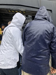 Several people wearing raincoats are standing closely together, with their backs to the camera. The scene appears to be outdoors with a wet environment, as indicated by water droplets on the clothing. The individuals are gathered near an entrance or passageway that has a warning sign saying 'Mind Your Head'.