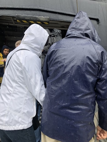 Several people wearing raincoats are standing closely together, with their backs to the camera. The scene appears to be outdoors with a wet environment, as indicated by water droplets on the clothing. The individuals are gathered near an entrance or passageway that has a warning sign saying 'Mind Your Head'.
