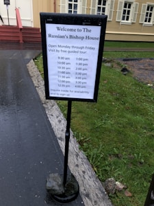 A signboard with the heading 'Welcome to The Russian's Bishop House' is displayed in front of a building with beige walls and white window shutters. The signboard lists the free guided tour times from 9:30 am to 4:00 pm, Monday through Friday. It is set on a path next to a grassy area.