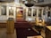 An ornately decorated interior of what appears to be a church with religious icons and paintings on the wall. There is a large, elaborate chandelier, lit candles on gold stands, and a carpeted platform leading to an altar with a gold decorative screen and a wooden table covered in a yellow cloth. The walls and doors are adorned with religious images and gold trim.