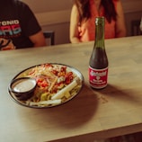 A wooden table with a colorful salad in a bowl, accompanied by a small dish of dressing. Next to the salad is a bottle of Señorial Sangria soda. The setting includes two people partially visible, seated around the table.