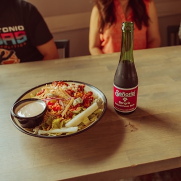 A wooden table with a colorful salad in a bowl, accompanied by a small dish of dressing. Next to the salad is a bottle of Señorial Sangria soda. The setting includes two people partially visible, seated around the table.