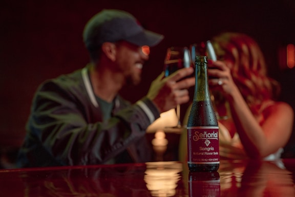 A bottle of Señorial Sangria, a natural flavor soda, is prominently placed on a table in a dimly lit setting. In the background, a blurred couple is clinking glasses, suggesting a celebratory or intimate atmosphere.