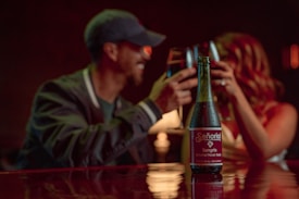A bottle of Se&ntilde;orial Sangria, a natural flavor soda, is prominently placed on a table in a dimly lit setting. In the background, a blurred couple is clinking glasses, suggesting a celebratory or intimate atmosphere.