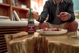 A person sits at a wooden table, holding a bottle of sangria-flavored soda in one hand and a small piece of food in the other. A plate with various appetizers, including cured meats and cheese, is placed on the table. Bookshelves and a sofa are visible in the background.