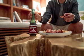 A person sits at a wooden table, holding a bottle of sangria-flavored soda in one hand and a small piece of food in the other. A plate with various appetizers, including cured meats and cheese, is placed on the table. Bookshelves and a sofa are visible in the background.