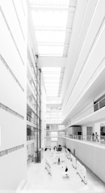 A modern, multi-story atrium with white walls and large windows. The space is filled with natural light from skylights above. People are present on different levels, and there are display boards and tables set up on the ground floor.